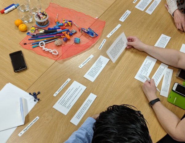 The image shows several people seated around a table, actively participating in an activity that involves arranging strips of paper with text. Various items, such as pens, small toys, and craft materials, are placed in the centre of the table, suggesting a creative or interactive group exercise.