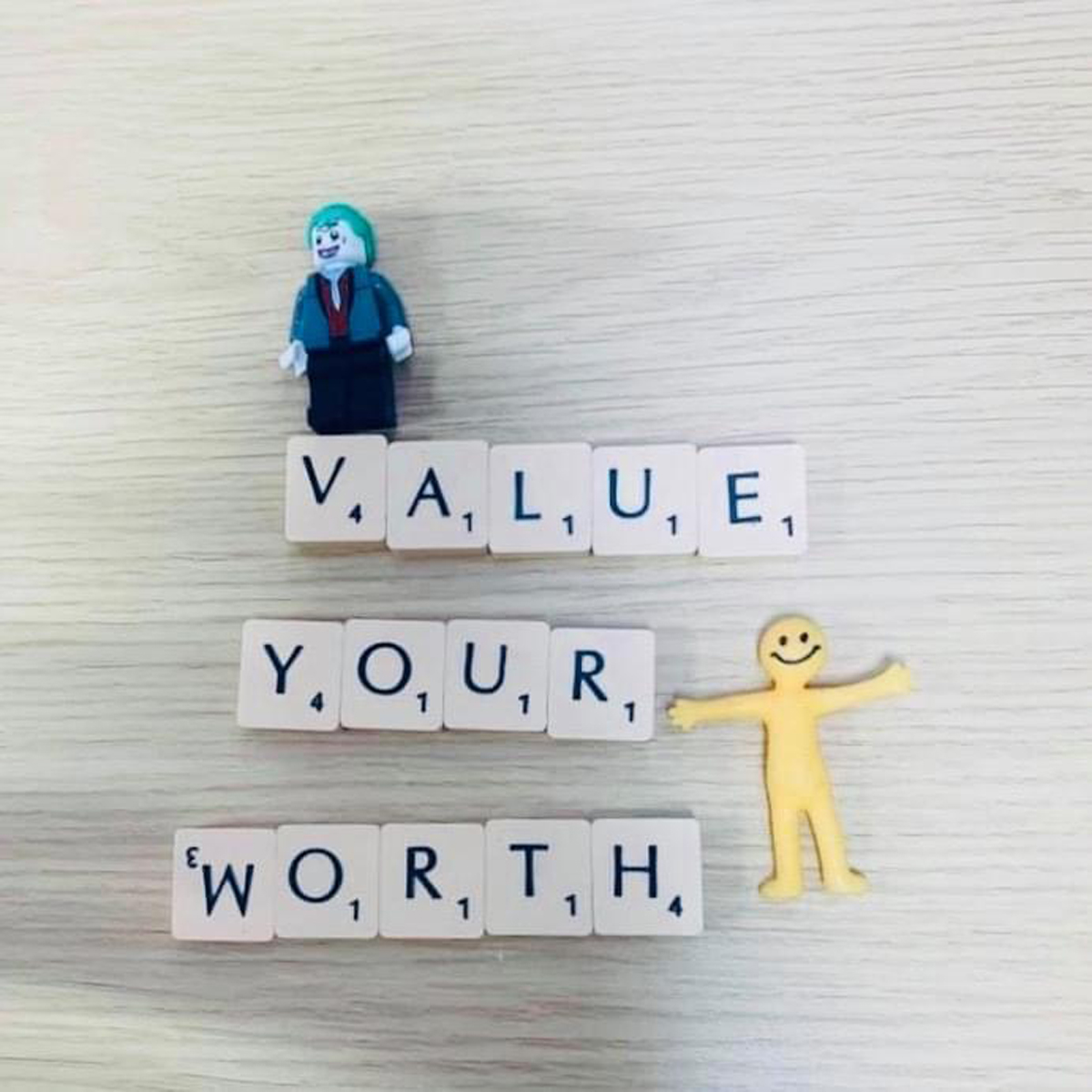 The image features the phrase "Value Your Worth" spelled out using Scrabble-style letter tiles. There is a small figurine of a person in a suit above the word "Value" and a yellow, smiley-faced figure with arms outstretched next to the word "Worth," symbolising the importance of self-worth and recognition.