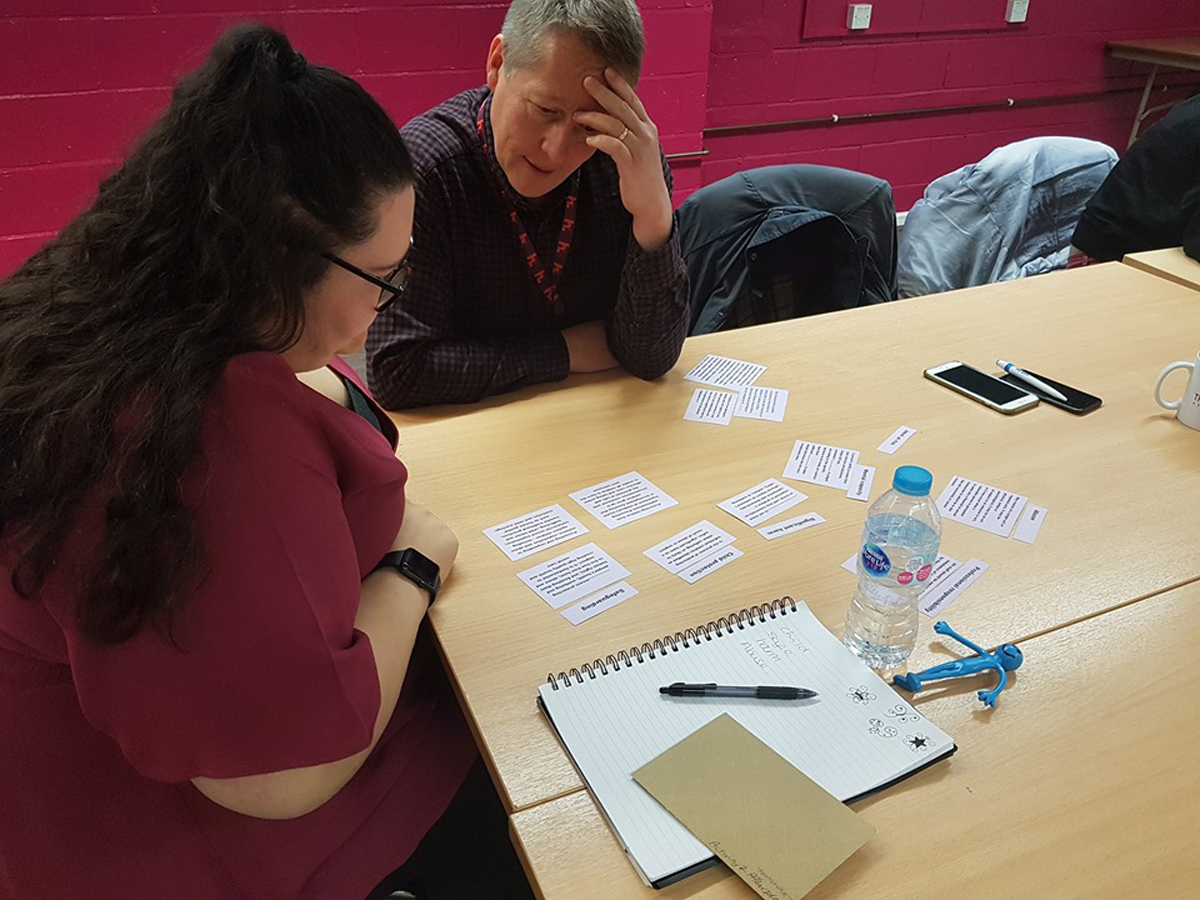 The image shows two people seated at a table, engaged in an activity involving arranging cards with text on them. There are also notebooks, a water bottle, and other materials on the table, indicating a collaborative and hands-on learning exercise.