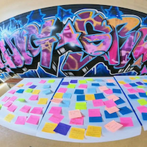 A table is covered with colourful sticky notes arranged in rows, set against a backdrop of vibrant graffiti art. The caption "Be Creative" suggests a focus on brainstorming, idea generation and collaborative creative activities in an inspiring environment.
