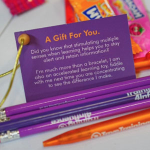 The image shows a small promotional gift pack, including colourful pens and a card with the message "A Gift For You." The card mentions the benefits of stimulating multiple senses for better learning. The caption "Develop Marketing" suggests a focus on creating engaging and effective marketing materials.