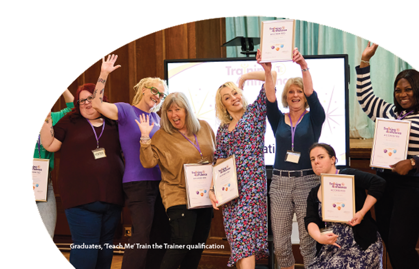 A group graduates are standing together, smiling, and holding thier certificates, celebrating a fantastic achievement. Some are striking playful poses with arms raised. They are in a bright room with a large screen in the background, creating a lively and joyful atmosphere.