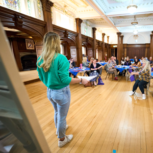 A person is standing at the front of a room, holding papers and speaking to a group seated around tables.