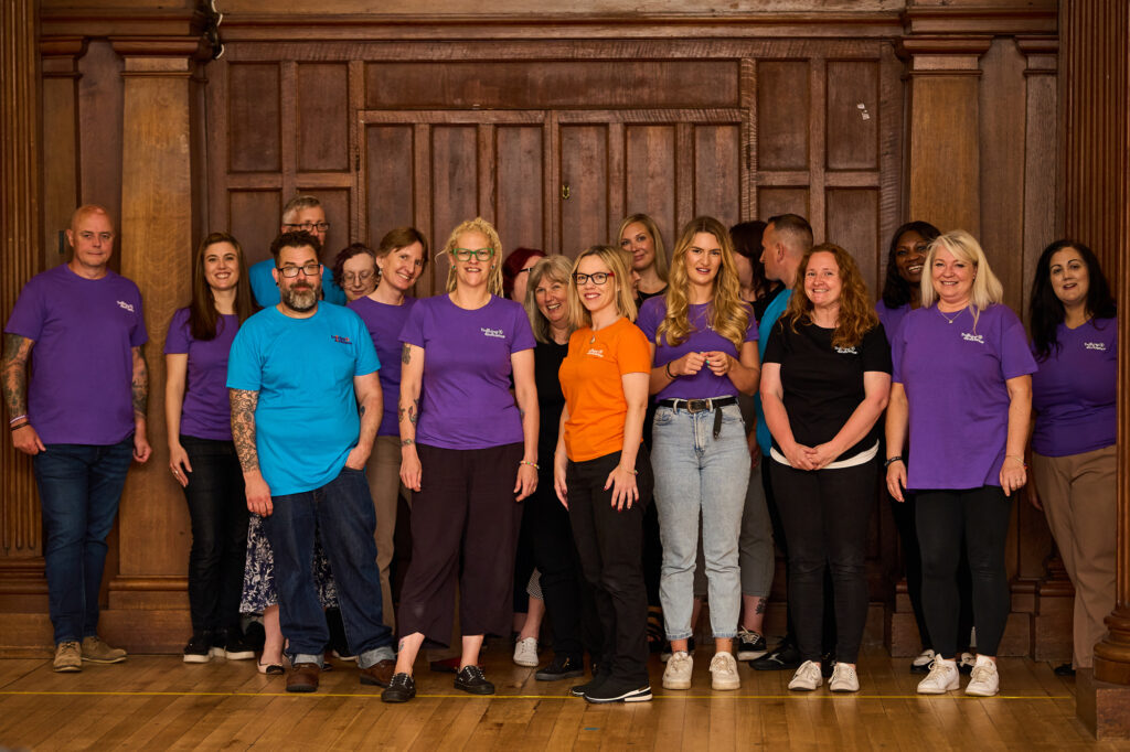 18 members of the Transform your Training team are standing together in a room with a wooden-panelled backdrop. They are casually dressed, many wearing brightly colored shirts with TAYE logo on, they are all smiling and laughing at the camera. The atmosphere is relaxed and happy.