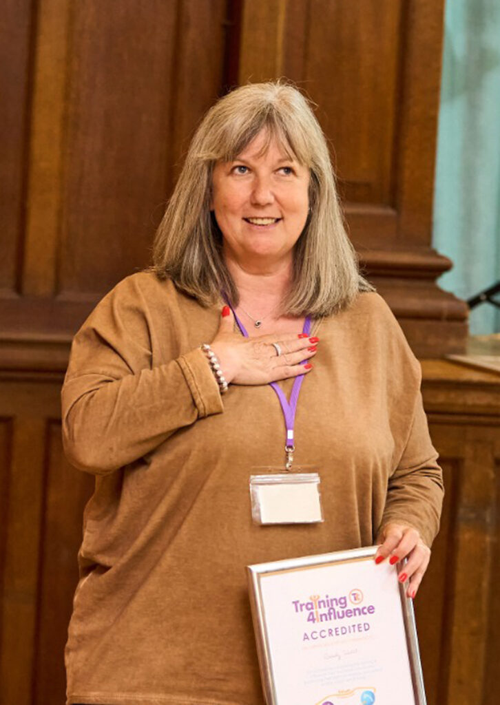 The image shows a Transform your Training graduate with shoulder-length grey hair, smiling and holding a certificate she has achieved. She is standing in a wooden-panelled room, with one hand on her chest, suggesting a moment of gratitude and recognition.