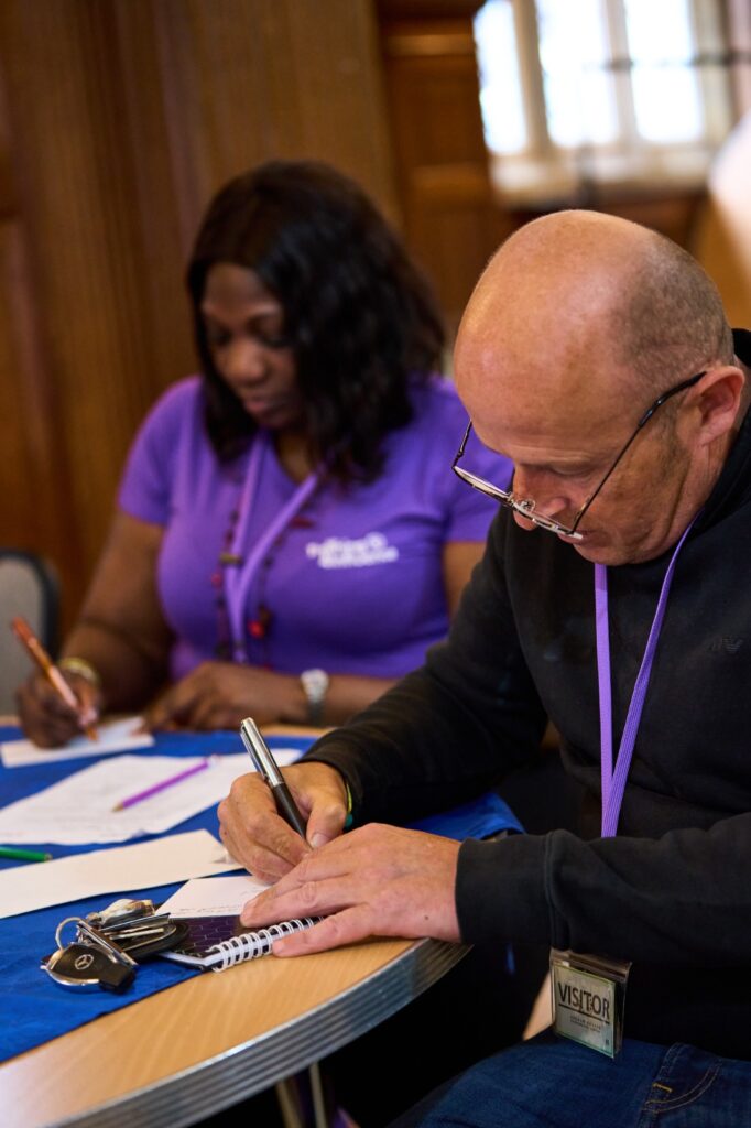 The image shows two training participants sitting at a table, both focused on writing. The man in the foreground is bald and wearing glasses, while the woman behind him, dressed in a purple t-shirt, is also writing, with a relaxed and studious atmosphere around them.