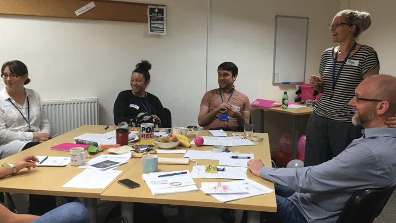 A small group of delegates are seated around a table in workshop setting. Papers, notebooks, and small toys are scattered on the table, and the group are engaged in a relaxed discussion.