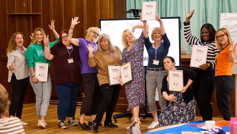 A group of graduates from the Train a Trainer Programme are standing together in a room, smiling and holding certificates of achievement. They are excited and are posing enthusiastically, celebrating their accomplishments.