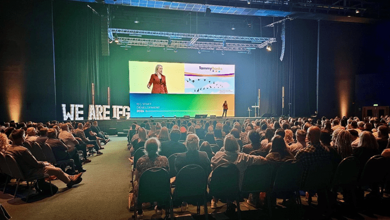 A large audience is seated in front of a stage where Tammy Banks .in a red jacket is presenting. A large screen behind the speaker displays colourful graphics related to the presentation.