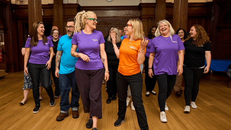 A group of Taye Facilitators are walking together in a spacious indoor setting, smiling and chatting. Many are wearing brightly coloured shirts with the TAYE logo on them. The atmosphere is casual and cheerful.