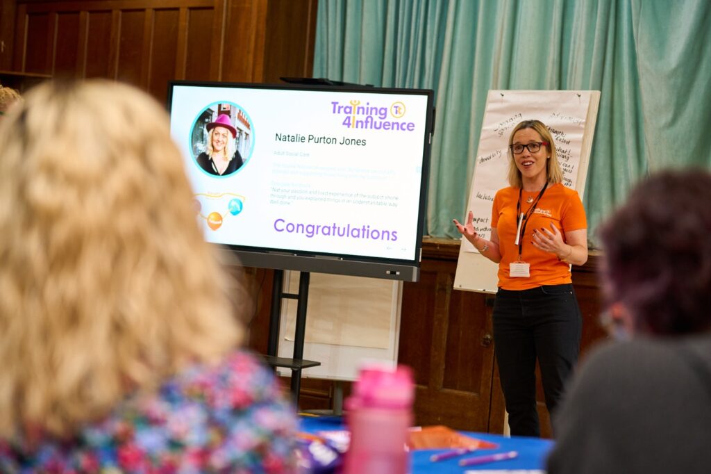 Tammy Banks is presenting at a Transform Your Training event, standing beside a screen displaying a congratulatory message. She is engaging with the audience, who are seated and listening attentively.