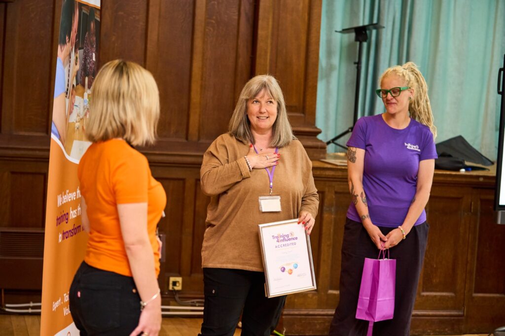 Tammy Banks and Faye Fox wearing colourful 'Training 4 Influence' T-shirts are smiling and watching a Transform your Training Graduate make a speech after receiving her award. The graduate is wearing a gold shirt and looks happy as she makes her speech while holding her certificate.