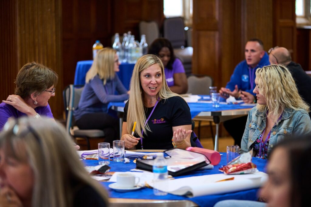 This image shows participants attending a face-to-face "Transform Your Training" event. A group of delegates are gathered in a room, seated and all participating in lively conversation with each other.