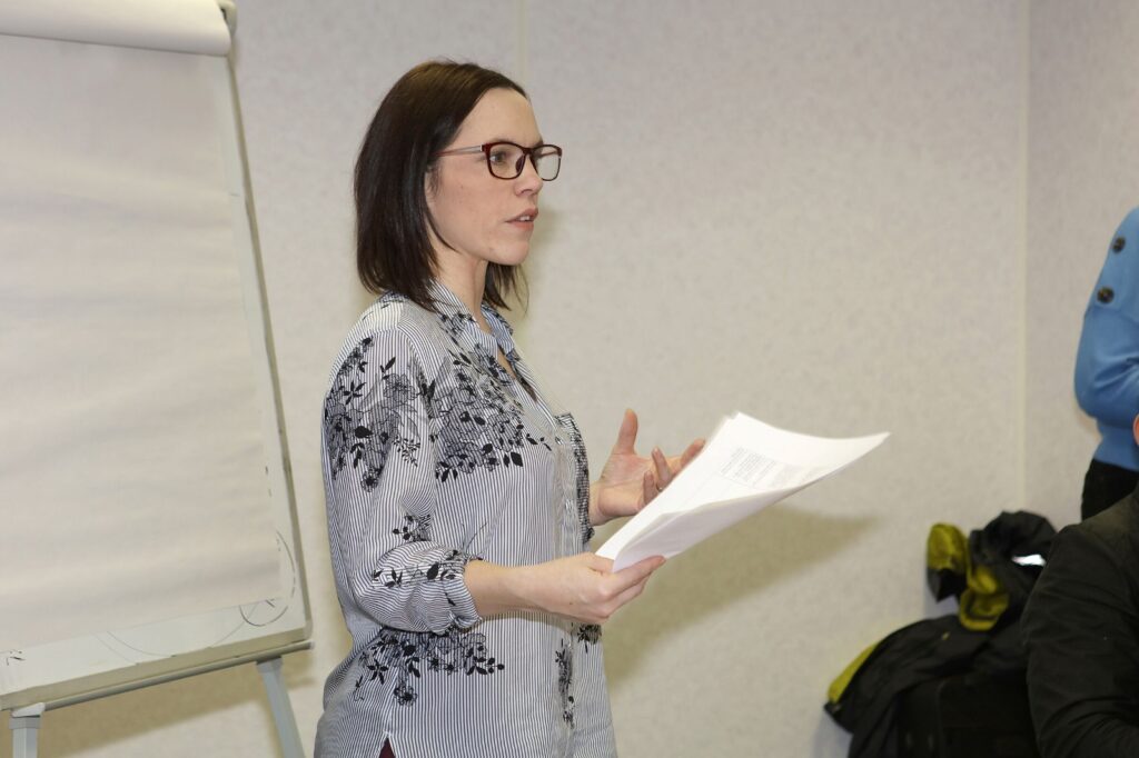 The image shows Tammy Banks with shoulder-length dark brown hair, holding a piece of paper. Behind her, there is a flip chart. Tammy is delivering training.