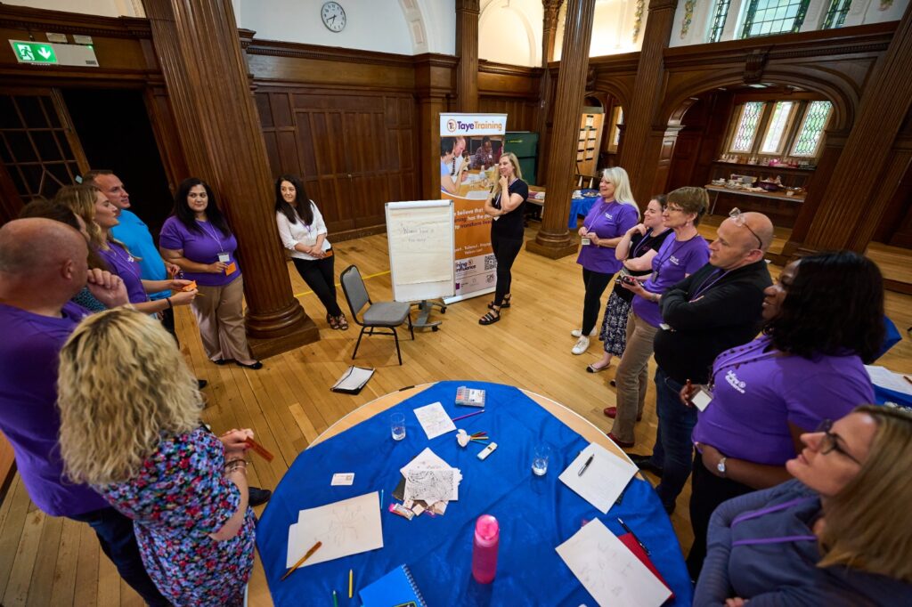 The image shows a Taye facilitator, addressing a seated group in a large room with wooden panelling. The audience is gathered around tables, listening attentively.