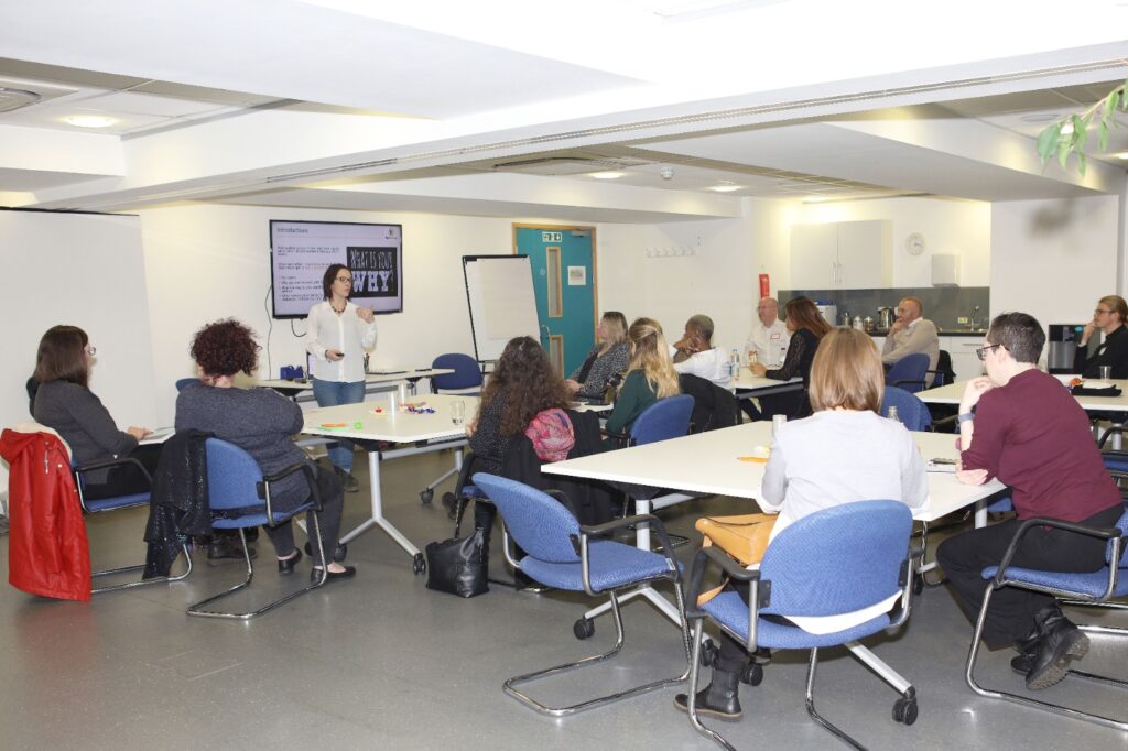 This image shows participants attending a face-to-face "Transform Your Training" event. A group of delegates are seated in a room all facing the Traner. who is delivering a presentation.