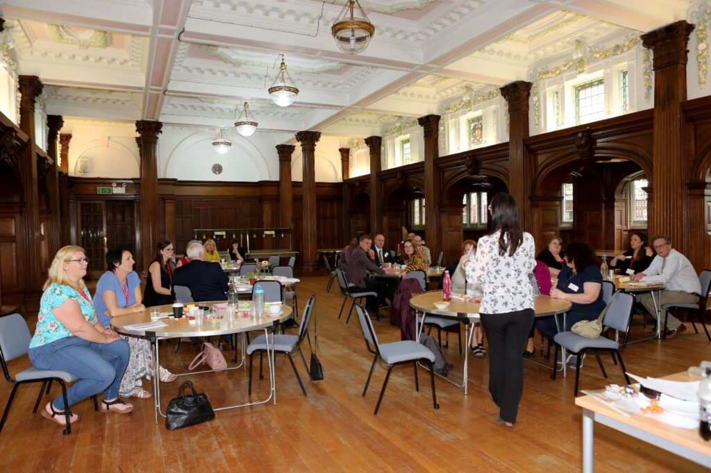 The image shows a Transform Your Training event taking place in a spacious, elegant room with high ceilings and ornate wood panelling. Several groups of people are seated around round tables, engaged in discussions or activities. A facilitator is standing near the centre of the room, possibly addressing participants or moving between groups.