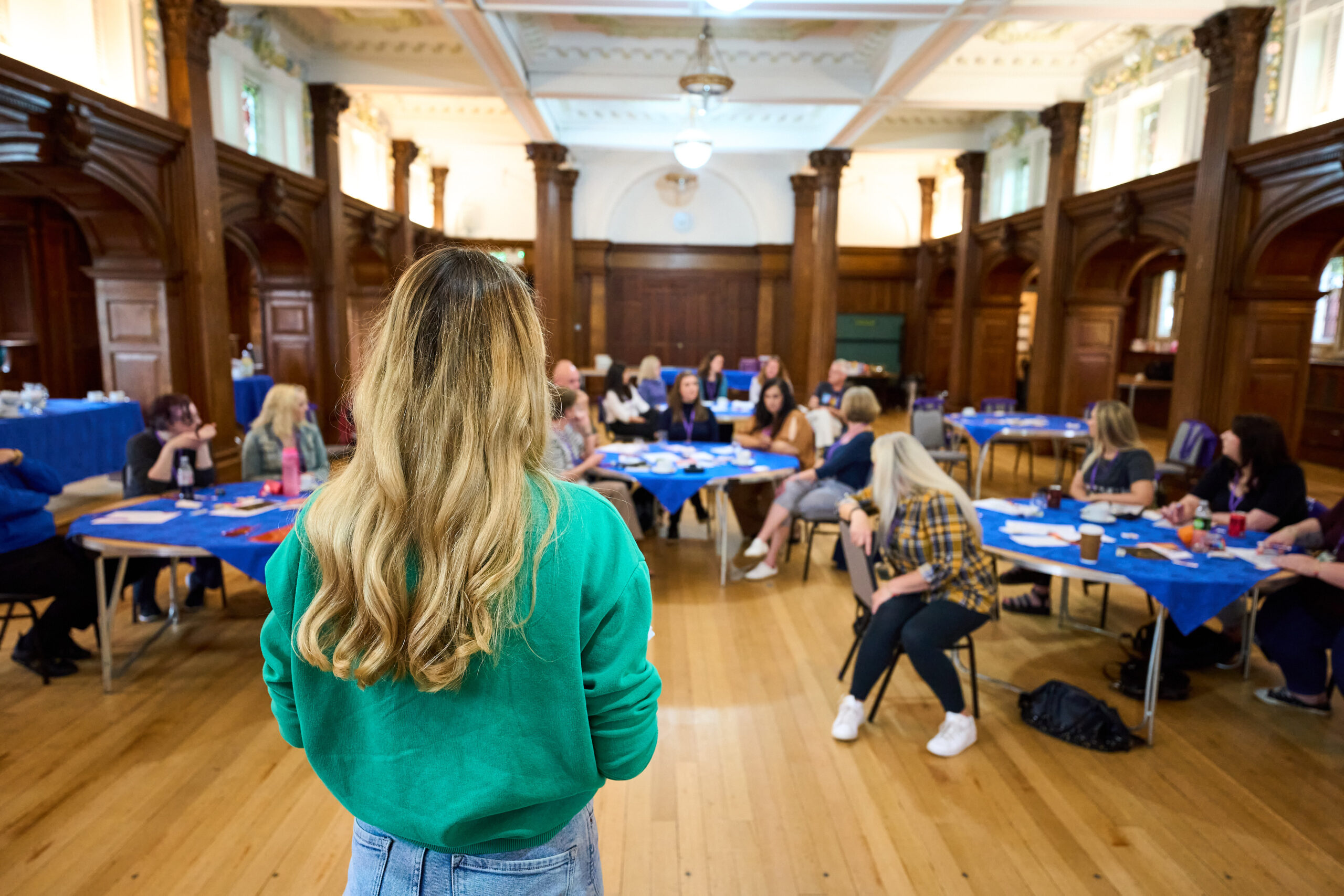 The image shows a woman with long blonde hair, wearing a green sweater, facing a group of people seated at tables in a spacious room with wooden walls and high ceilings. The group appears to be engaged in a workshop or meeting, with the woman leading or presenting to the attendees.
