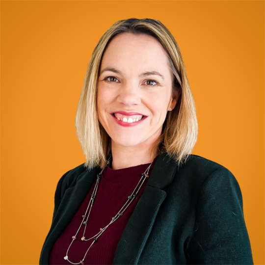 The image shows Tammy Banks with shoulder-length blonde hair, smiling and wearing a dark blazer over a maroon top. She is posed against a bright orange background, creating a professional and vibrant portrait.