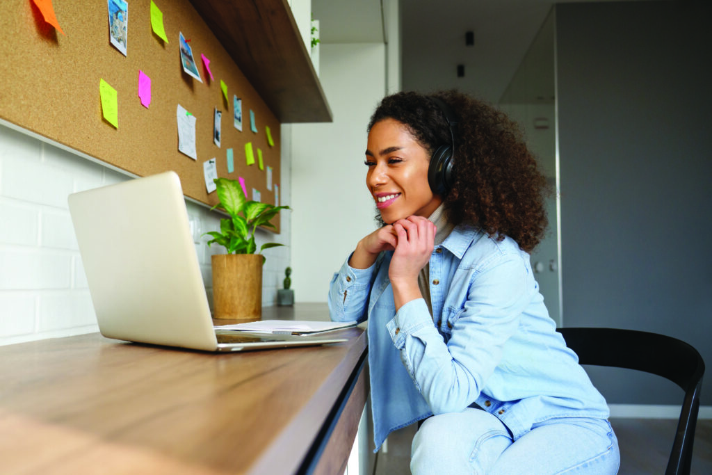 The image shows a woman sitting at a desk, smiling while looking at her laptop. She is wearing headphones and appears to be in a casual home office setting with a corkboard and colourful sticky notes on the wall behind her.