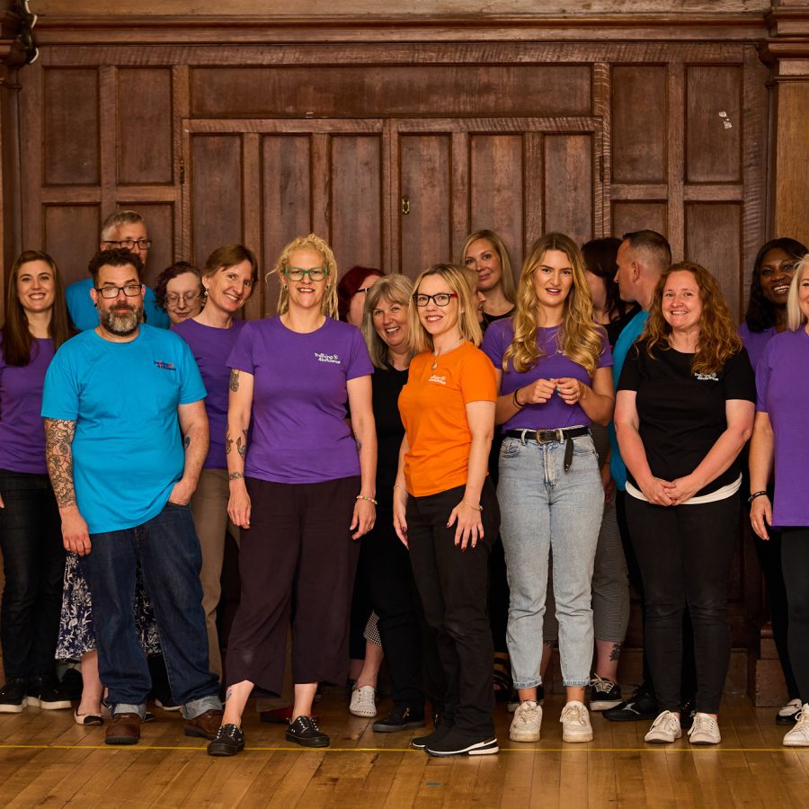 18 members of the Transform your Training team are standing together in a room with a wooden-panelled backdrop. They are casually dressed, many wearing brightly colored shirts with TAYE logo on, they are all smiling and laughing at the camera. The atmosphere is relaxed and happy.