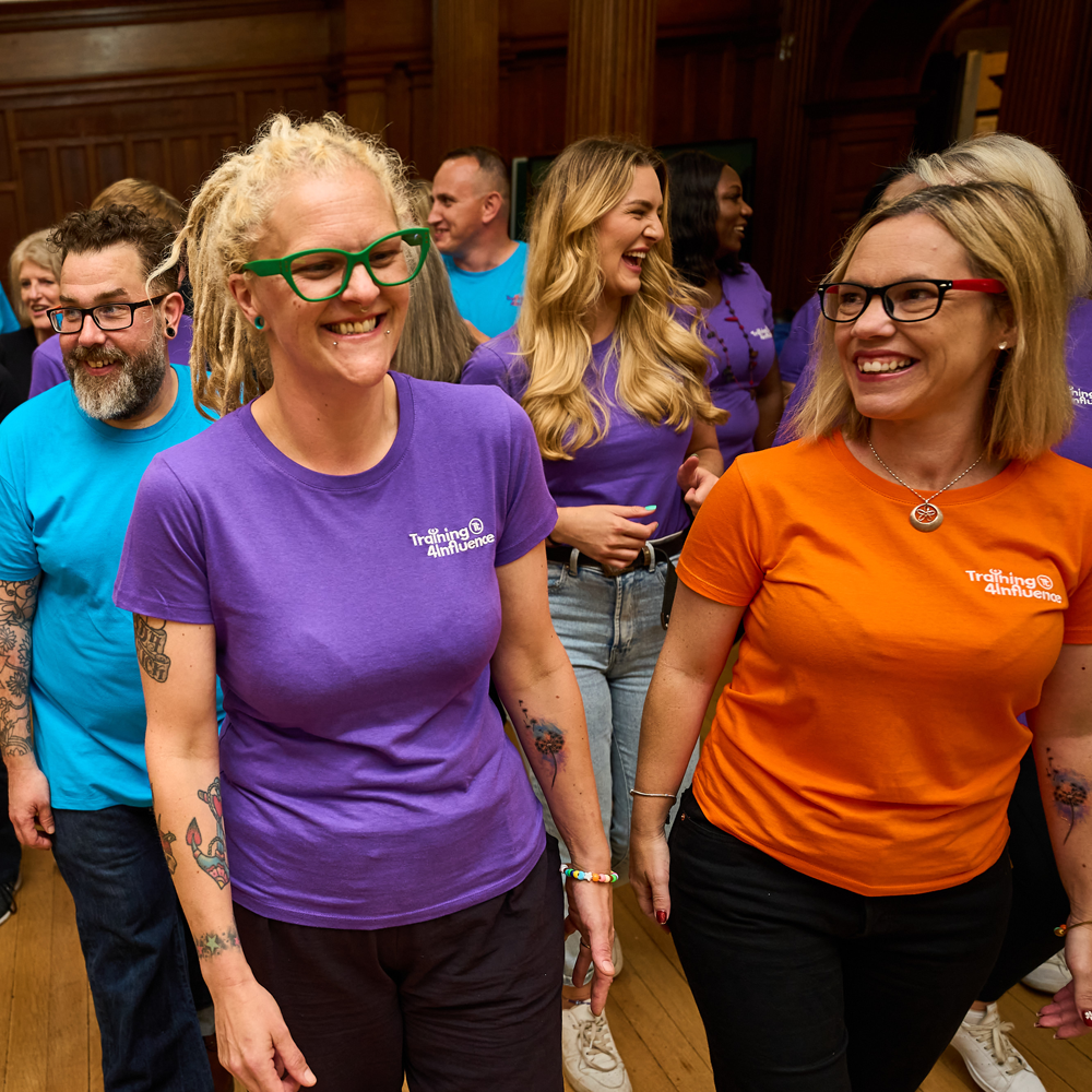 Tammy Banks and Faye Fox wearing colourful 'Training 4 Influence' T-shirts are smiling and walking together in a room, followed by the Transform your Training team. They are enjoying a lively positive atmosphere.