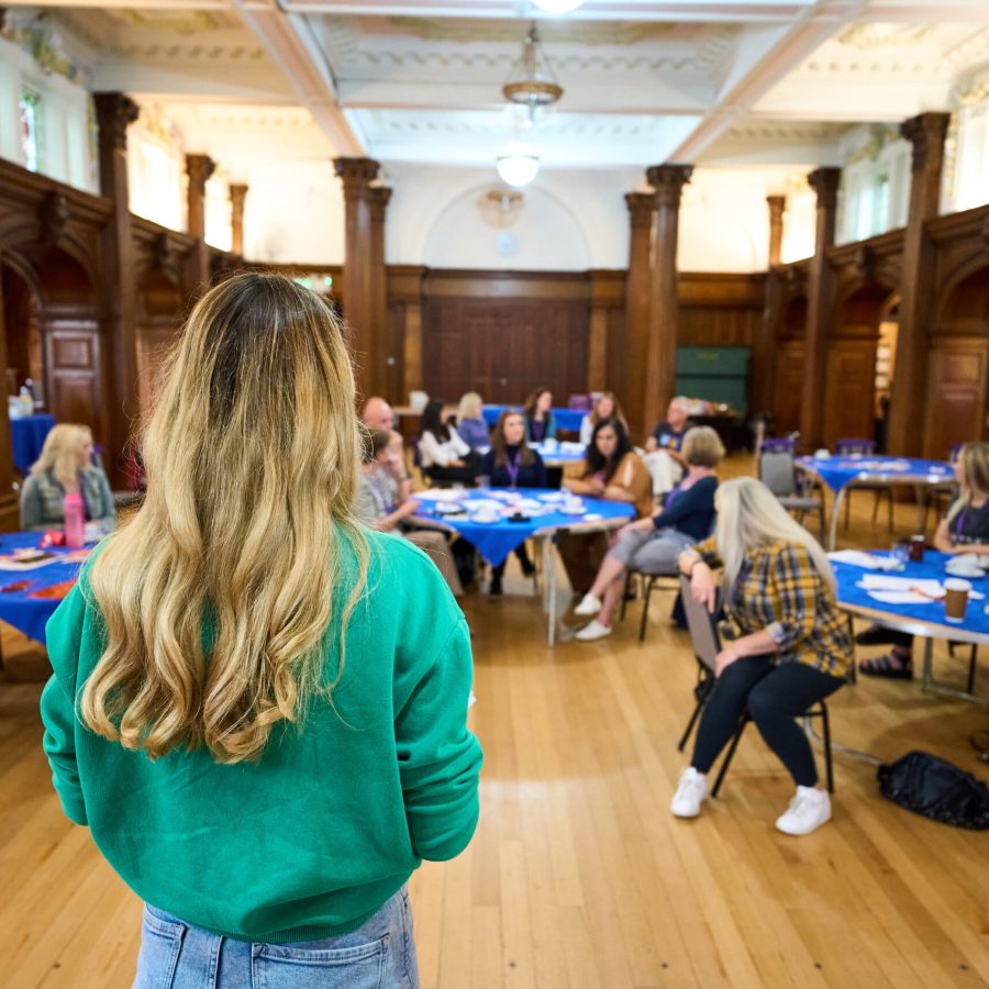 The image shows a woman with long blonde hair, wearing a green sweater, facing a group of people seated at tables in a spacious room with wooden walls and high ceilings. The group appears to be engaged in a workshop or meeting, with the woman leading or presenting to the attendees.