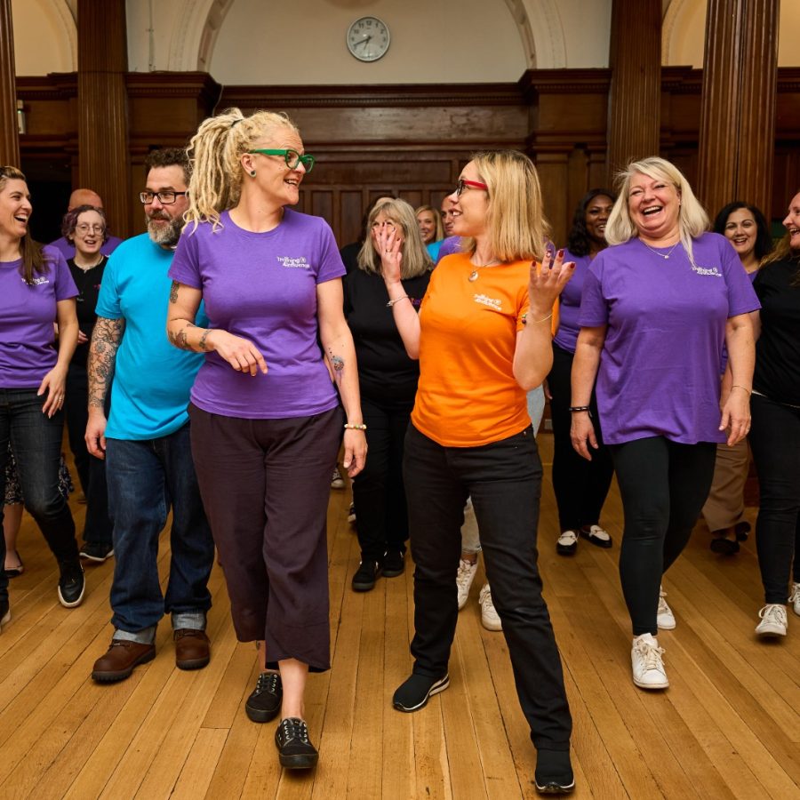 The image shows the Transform you Training team walking together in a large room, smiling and chatting. Tammy Banks and Faye Fox are in the foreground, Tammy wearing an orange t-shirt and Faye in a purple t-shirt, they are leading the group, creating a friendly and lively atmosphere.