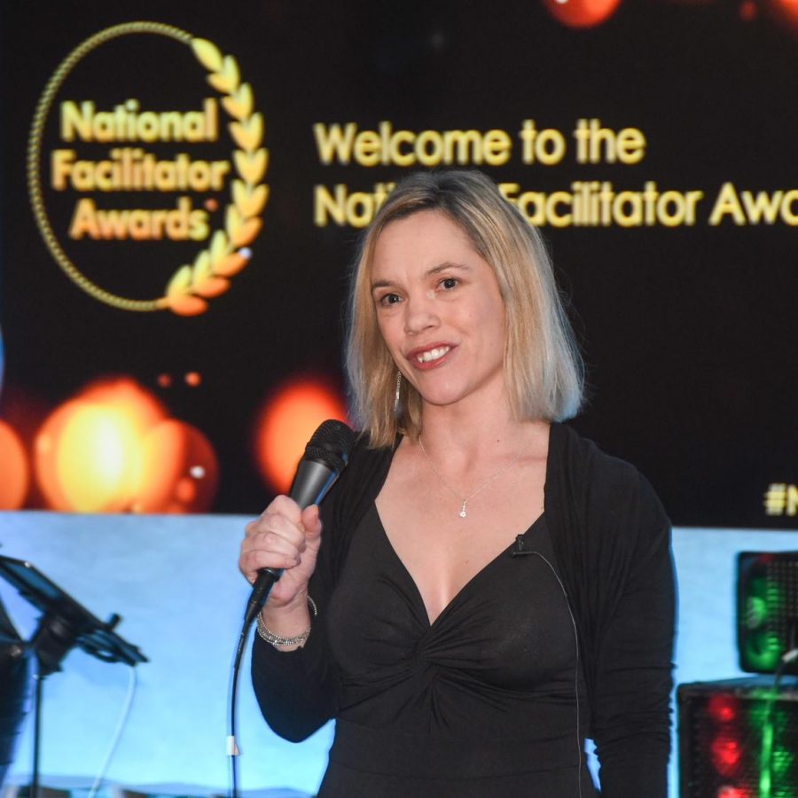 The image shows Tammy Banks with shoulder-length blonde hair, holding a microphone and speaking at the National Facilitator Awards. Behind her, there is a screen that reads "National Facilitator Awards" with a laurel logo, she is presenting at the awards ceremony.