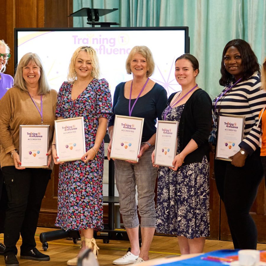 The image shows six women standing in a line, smiling and holding certificates. They are in a casual, celebratory setting with a screen in the background, marking their achievement