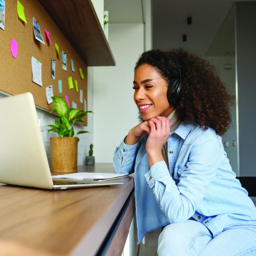 The image shows a woman sitting at a desk, smiling while looking at her laptop. She is wearing headphones and appears to be in a casual home office setting with a corkboard and colourful sticky notes on the wall behind her.