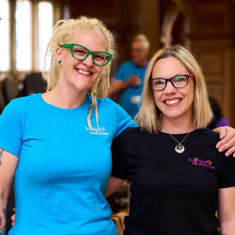 Tammy and Faye are smiling and standing side by side, each with an arm around the other. Faye is wearing a bright blue t-shirt and Tammy a black t-shirt, both have the words TAYE printed on them.